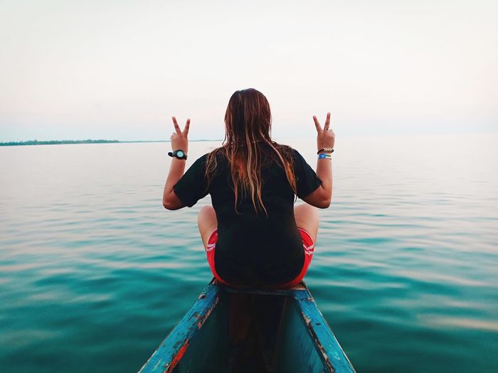 Rear view of woman showing peace sign on boat in sea against sky