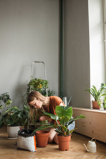Woman pours soil into pot kneeling surrounded by plants at home