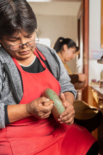Concentrated peruvian female artisan in apron painting ceramic bowl with green color on stand while working in light professional workshop