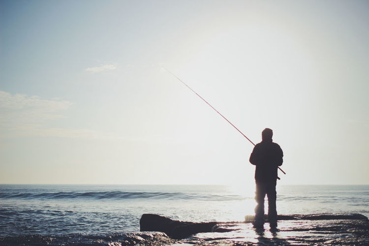 Rear view of man fishing in sea against sky on sunny day
