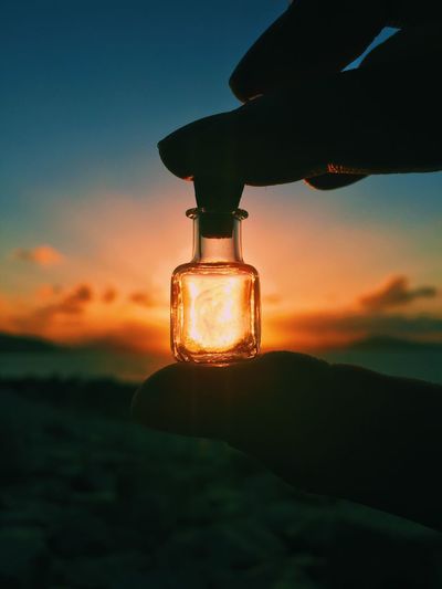 Cropped hand holding small jar at beach against sun during sunset