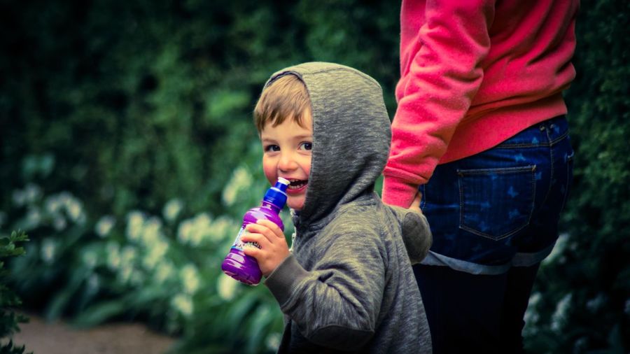 Portrait of happy boy holding water bottle while walking with mother against plants