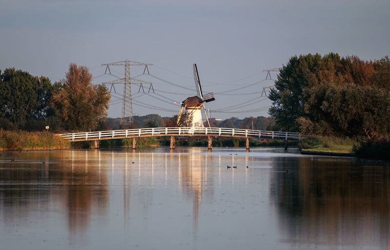 Scenic view of traditional windmill and bridge by river against sky