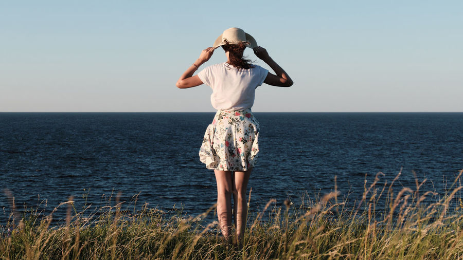 A woman with a hat stands on the shore. view from the back.