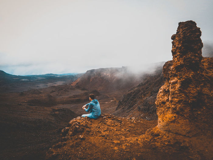 Man sitting on rock against sky during foggy weather