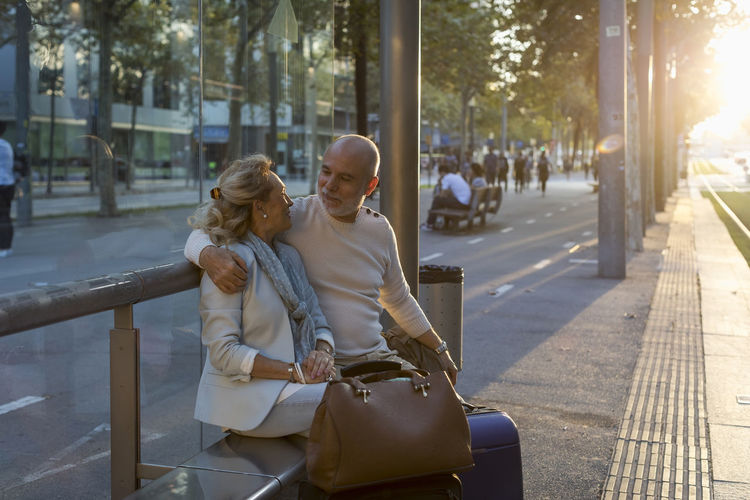 Spain, barcelona, senior couple with baggage sitting at tram stop in the city at sunset