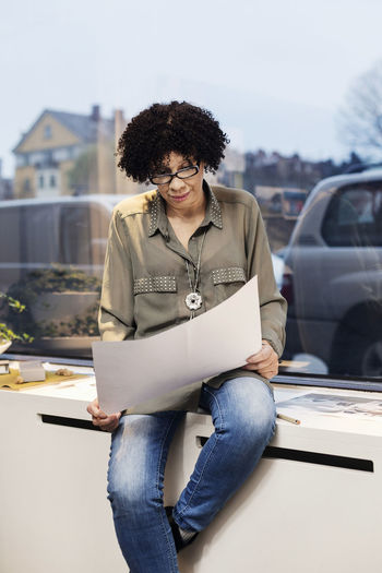 Businesswoman looking at blueprint while sitting on window sill