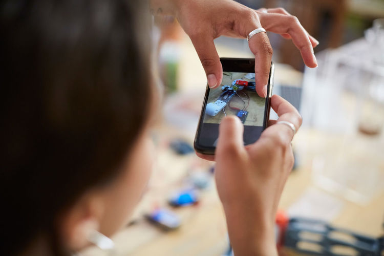 Cropped image of young female engineer touching mobile screen while photographing model at workshop