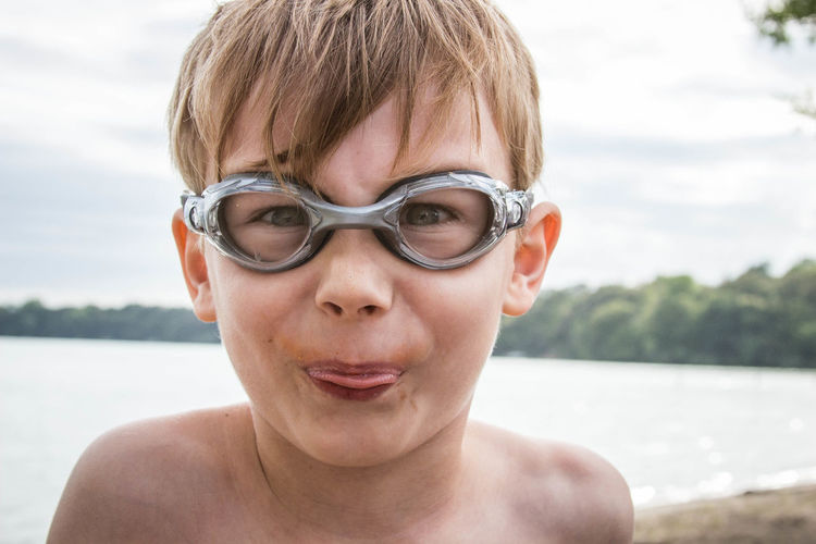 Close-up portrait of boy wearing swimming goggles while teasing against sky