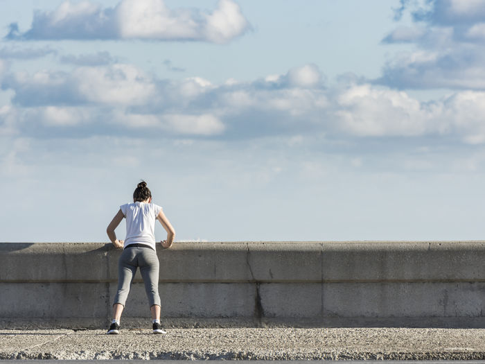 Rear view of woman exercising on seawall against sky
