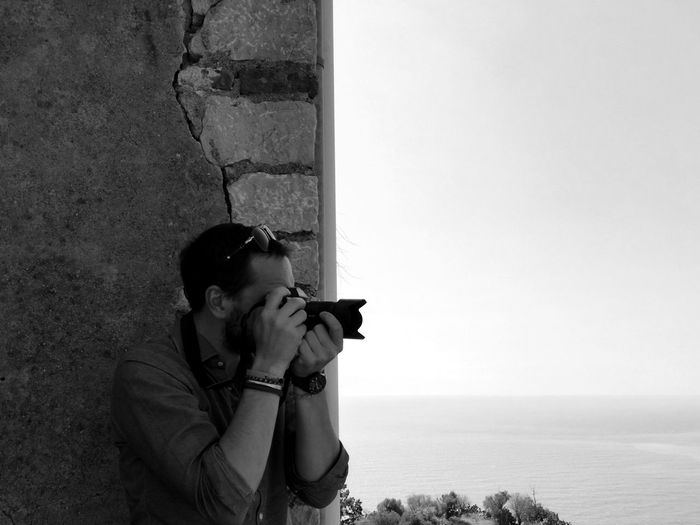 Man photographing through dslr while standing against wall by sea