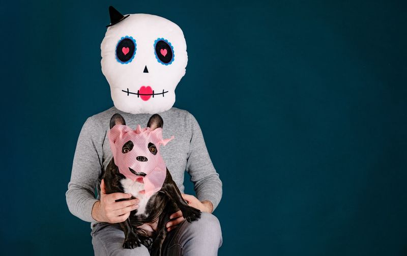 Man with halloween mask holding masked french budog dog against blue background