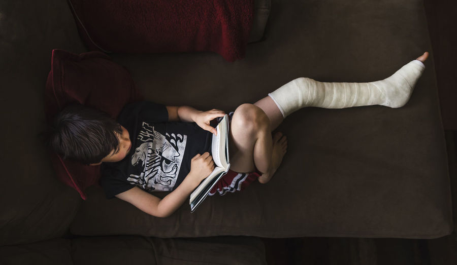 High angle view of boy with broken leg reading book while lying on sofa at home