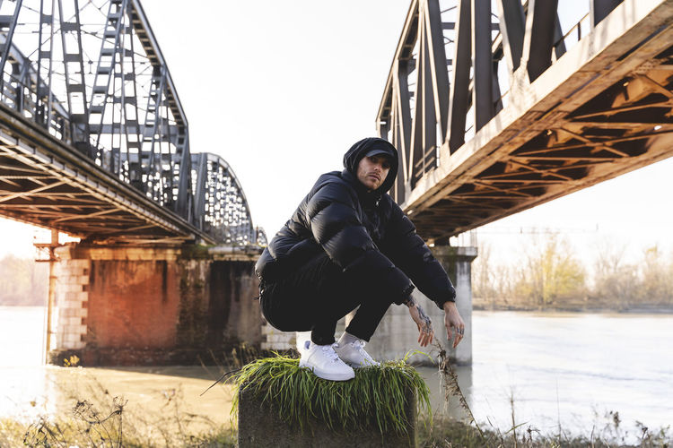 Portrait of young man crouching on land by bridges in city
