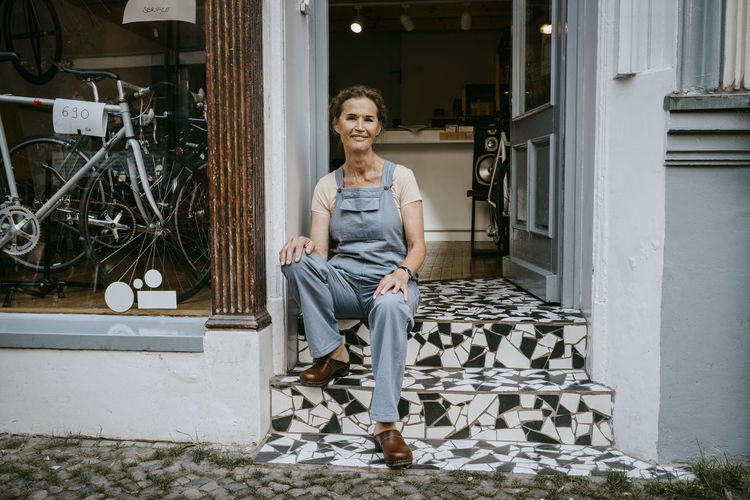 Portrait of confident senior owner sitting on steps of bicycle shop