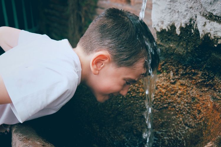 Side view of playful boy putting head under running water outdoors