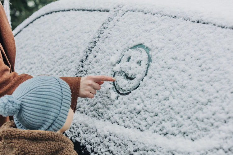 Boy standing by mother drawing human face on car window covered with snow