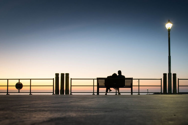 Silhouette friends sitting on bench against clear sky during sunset