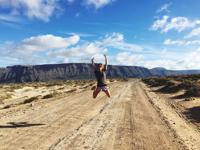 Happy woman jumping over dirt road by rocky mountains against sky