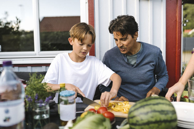 Son cutting vegetable sitting by father in balcony