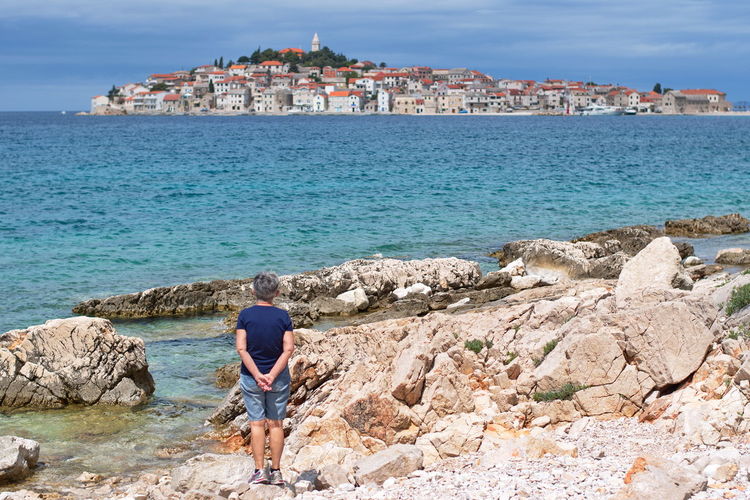 Senior woman standing on rocky beach looking at cityscape of mediterranean town over the sea