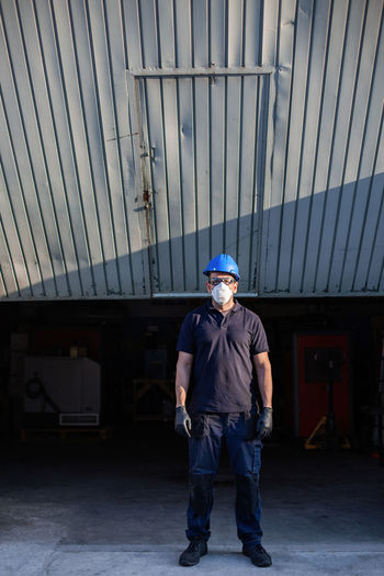 Adult male mechanic in protective glasses and respirator with blue hardhat looking at camera while standing near workshop building on street