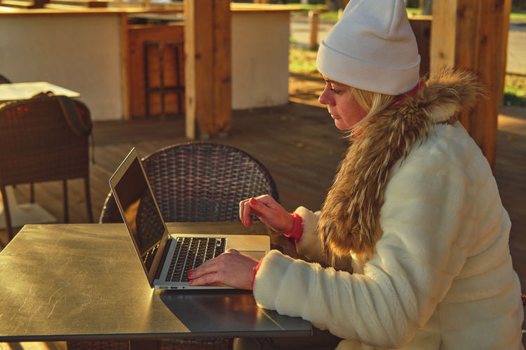 A woman in a sweater and a fur coat is sitting on the terrace of a cafe with a laptop.
