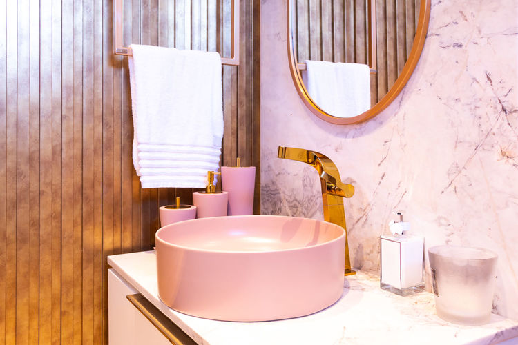 Interior of bathroom with towel on hanger near pink dispensers with toiletries near round washbasin with golden faucet under mirror