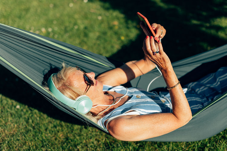 Caucasian senior lady swings in the hammock listening to music with phone and headphones.