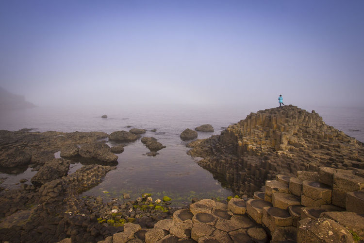 Mid distance view of woman standing on rocks by sea against sky during foggy weather