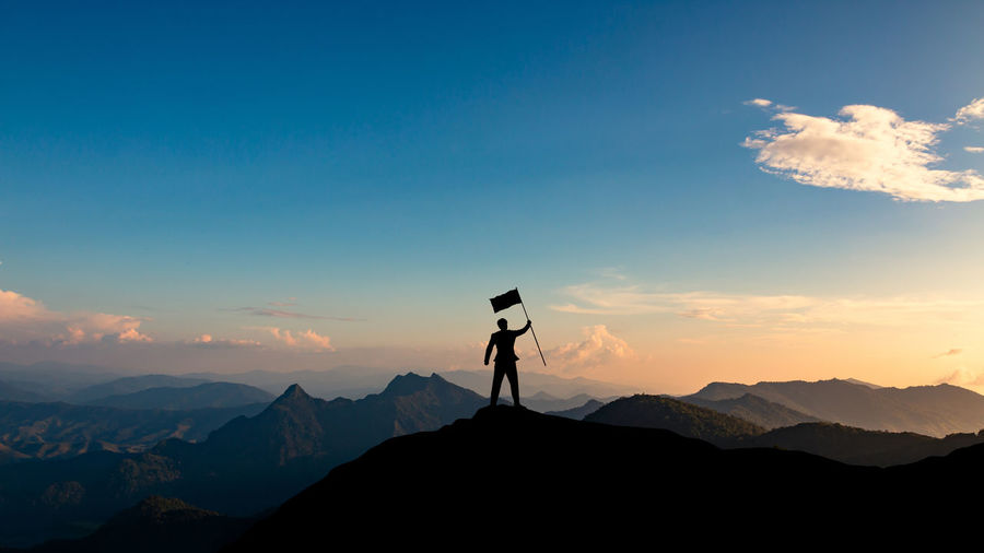 Silhouette man holding flag while standing on mountain peak against sky during sunset