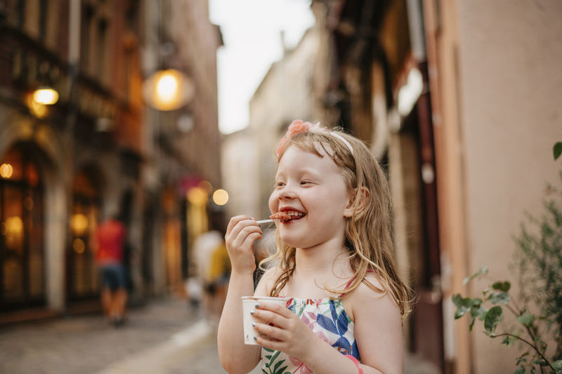 Happy girl eating food while standing on street against buildings in city