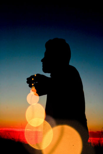 Silhouette man holding camera while sitting against sky during sunset