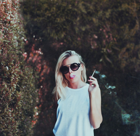 Portrait of mid adult woman wearing sunglasses while smoking against plants at park