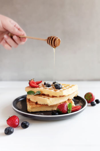 Hand holding honey spoon with a stream of honey over stack of waffles on a plate on the gray table