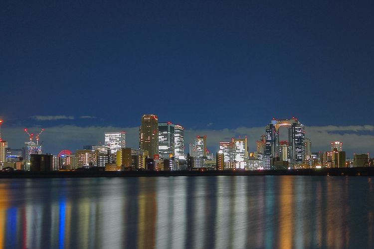 Illuminated city buildings by river against sky