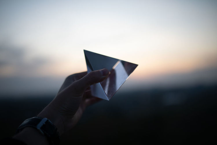 Cropped hand holding prism against sky during sunset
