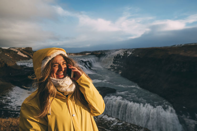 Pretty girl with yellow raincoat using mobile phone together a waterfall in iceland ..