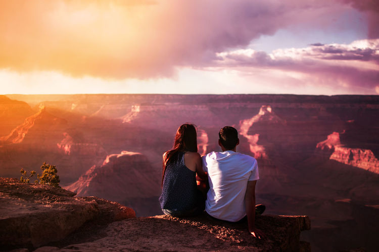Rear view of couple sitting on cliff against sky during sunset