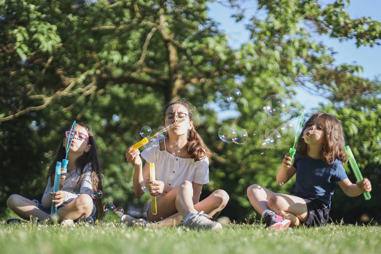 Portrait of three beautiful caucasian girl sisters blowing soap bubbles while sitting in the park