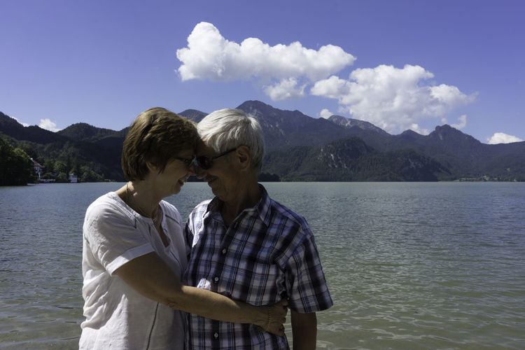 Happy senior couple embracing by lake against sky