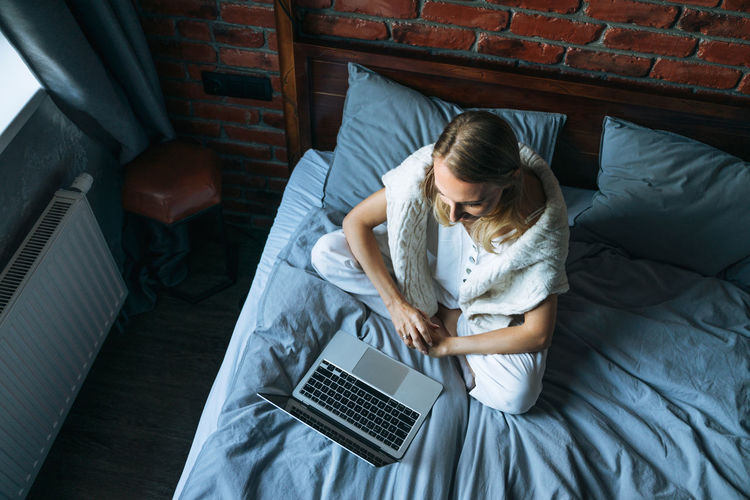 Adult woman freelancer with blonde long hair in casual clothes using laptop sitting on bed 