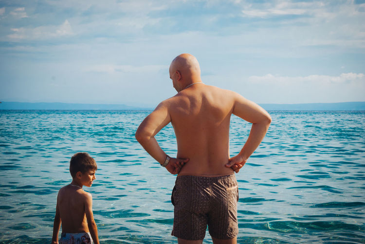 Rear view of shirtless father and son standing at beach against sky