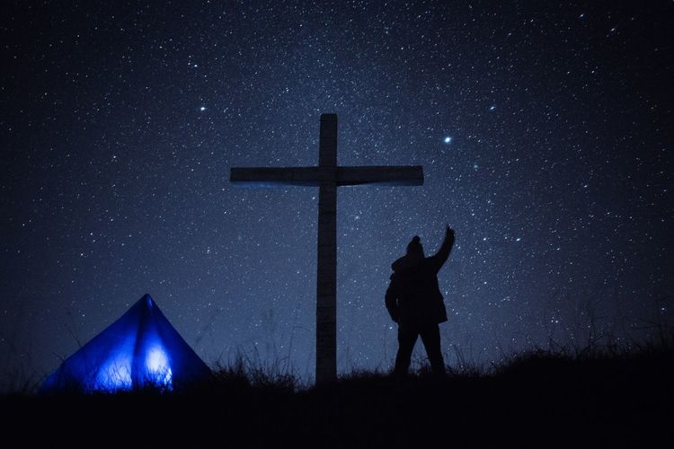 Silhouette person standing on cross against sky at night