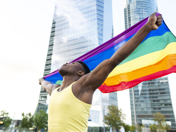 Young man wearing yellow vest holding rainbow flag in front of skyscrapers