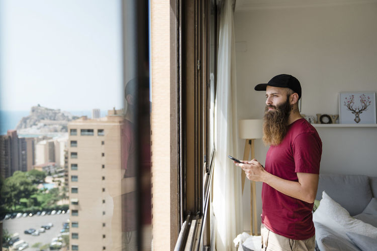 Side view of thoughtful bearded man looking through window while holding mobile phone at home
