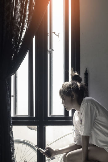 Side view of young woman looking through window at home