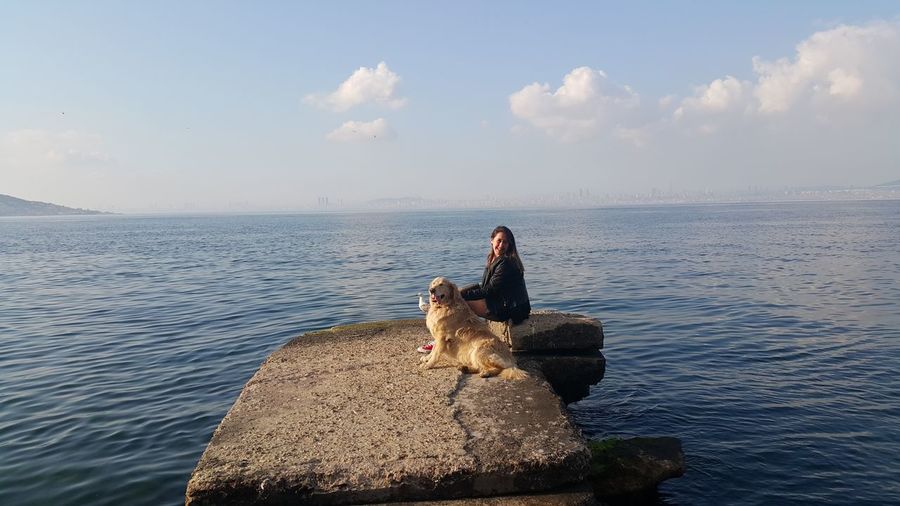 Portrait woman sitting with dog on rock by sea against sky