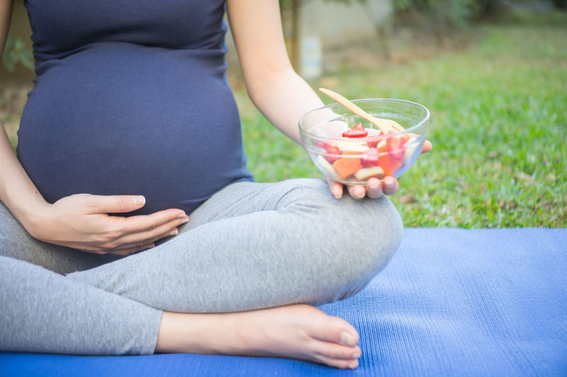 Low section of pregnant woman holding bowl with fruits while sitting on exercise mat