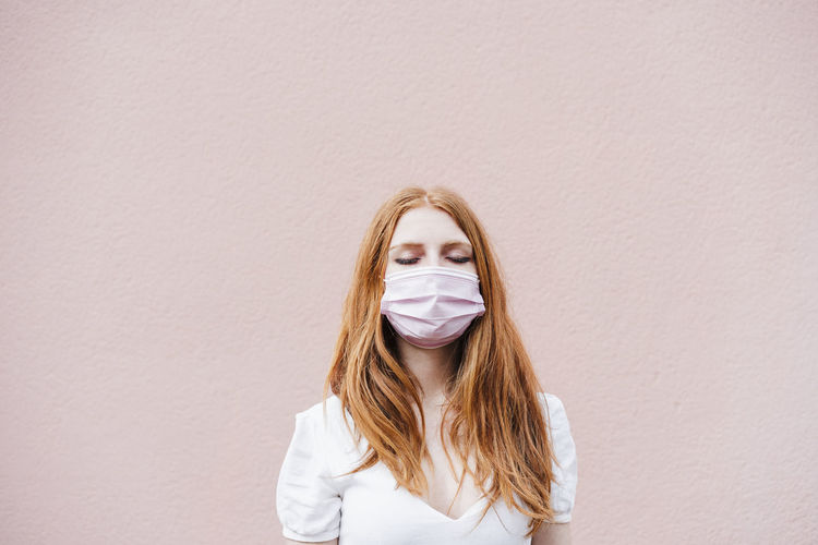 Young woman with eyes closed wearing protective face mask while standing in front of pink wall during covid-19